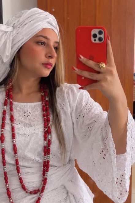 Mãe Lavínia de turbante e roupa branca, com guias vermelhas, tirando foto no espelho