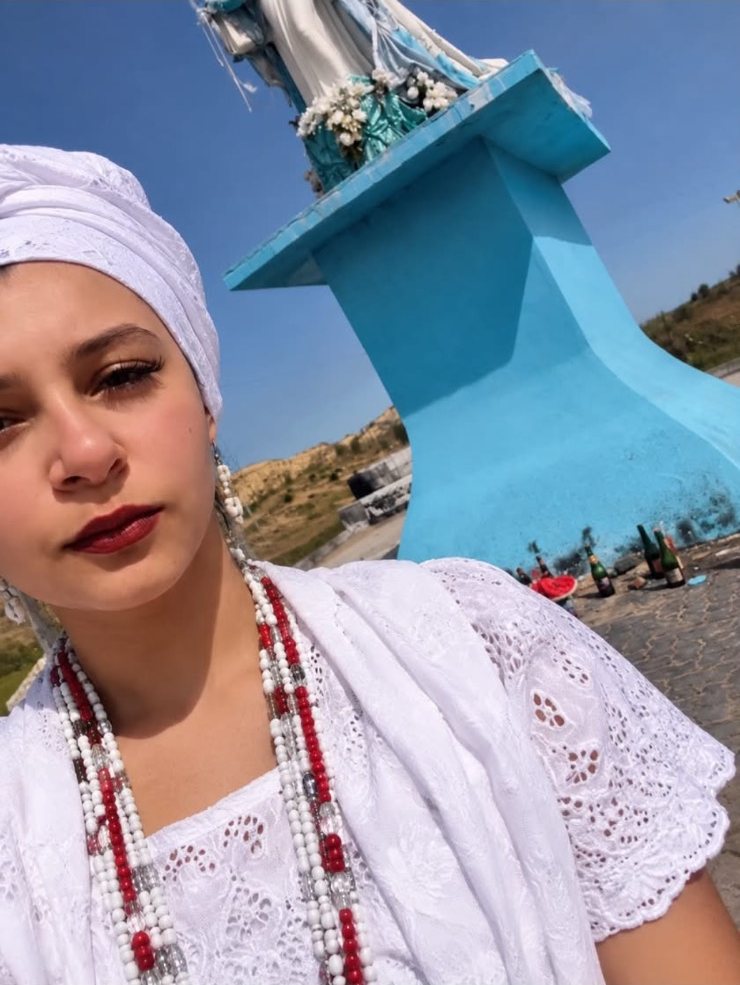 Mãe Lavínia de turbante branco e guias vermelhas e brancas, ao ar livre sob o céu azul